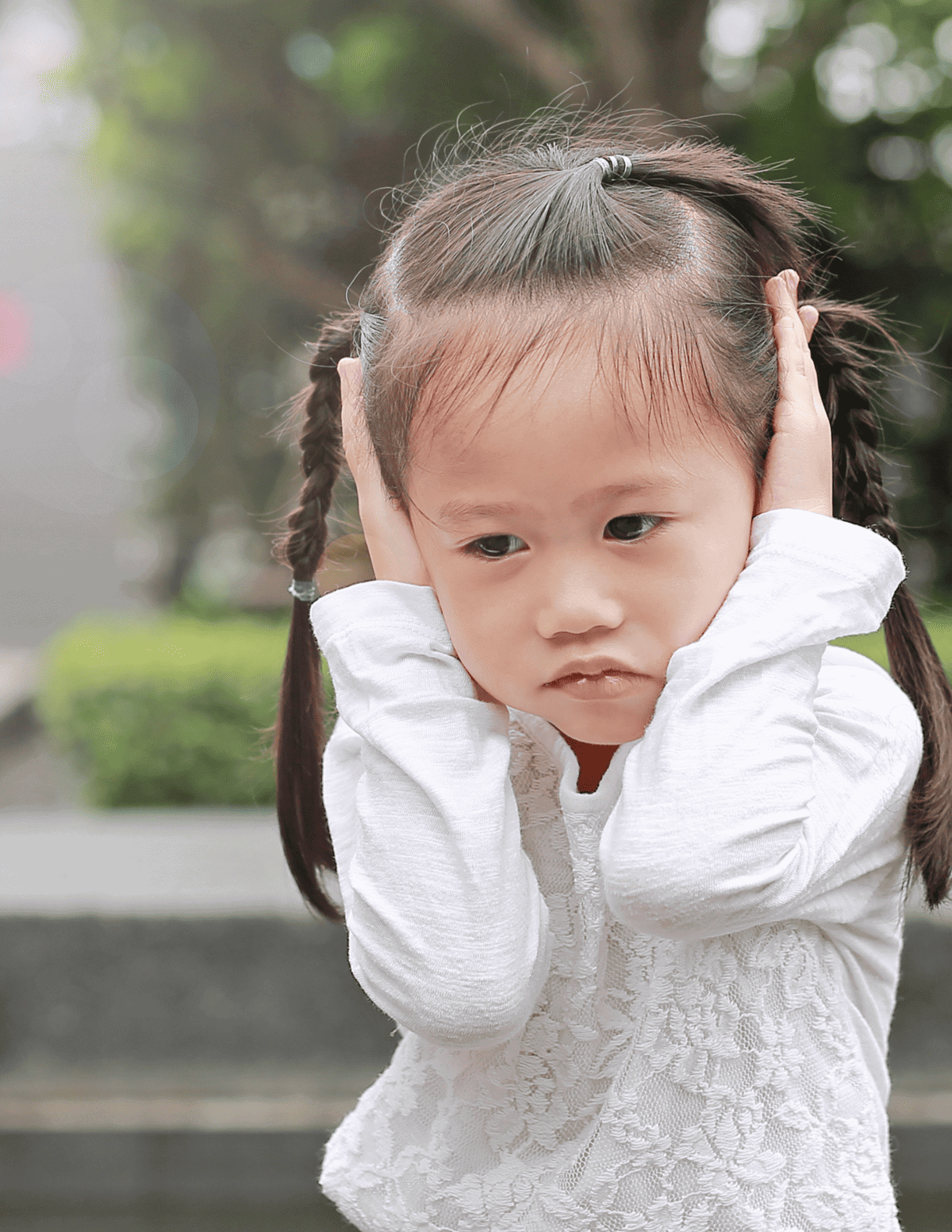 Preschooler covering both ears with each hand looking sad and at the ground.