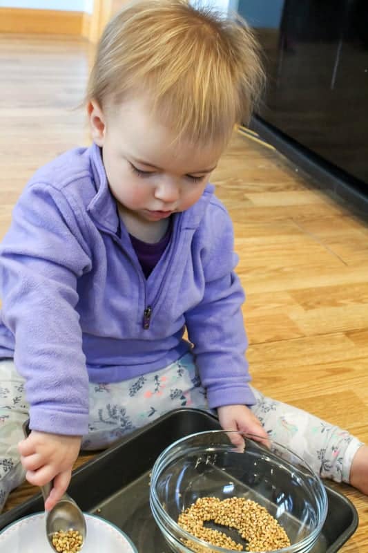 Toddler sitting on the floor transferring rice from one bowl to another with a spoon. 