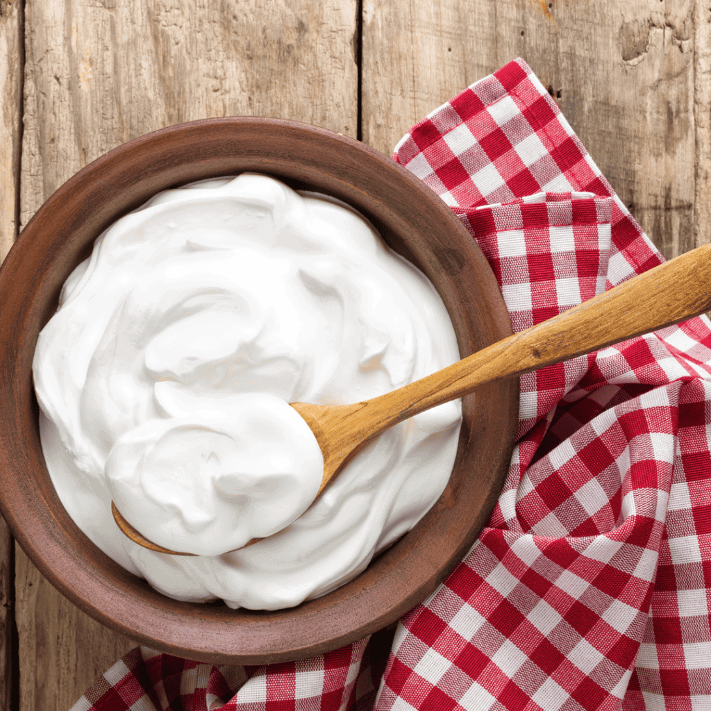 a wooden bowl of yogurt with a wooden spoonful of yogurt on top sitting on a rough wooden surface with a red and white checkered towel.