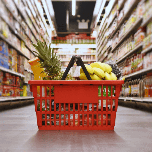 Red shopping basket filled with fresh food: bananas, pineapple, blueberries, potatoes, etc. sitting on the aisle way floor. The shelves are filled with juices and processed food snacks