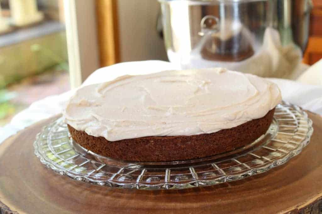 One layer of apple spice cake on the crystal cake plater with cinnamon buttercream frosting smoothed over the top. The platter is on a wood tree slice surrounded by white linen with the mixing bowl of frosting in the background. 