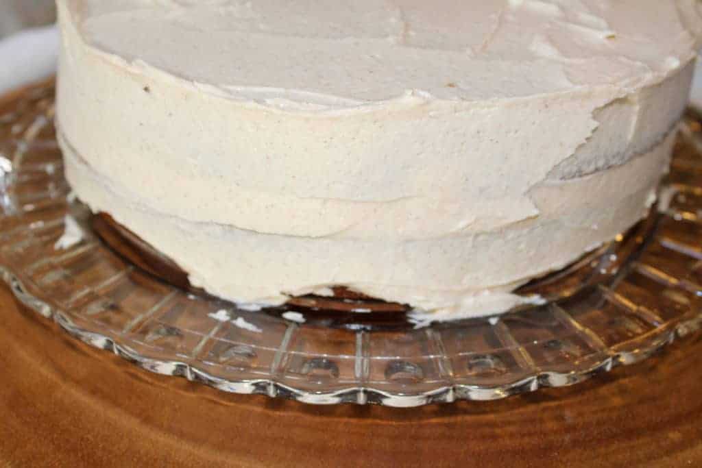 Apple spice cake with two layers with icing on the top and sides. It is sitting on a crystal platter on a wood tree slice. 