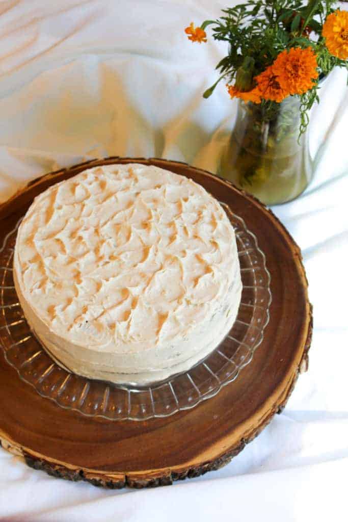 gluten and dairy free apple spice cake with cinnamon buttercream frosting on a glass cake platter sitting on a round wooden lazy susan. There is a white tablecloth underneath of it and a vase in the upper right hand corner with orange and yellow marigolds.