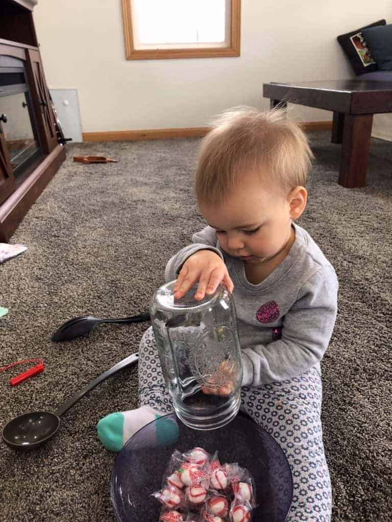 toddler girl wearing a sweater and leggings playing on the floor. She is dumping a mason jar filled with soft peppermint candies into a plastic bowl on the floor in front of her. A ladle and a serving spoon are on the floor behind her. 