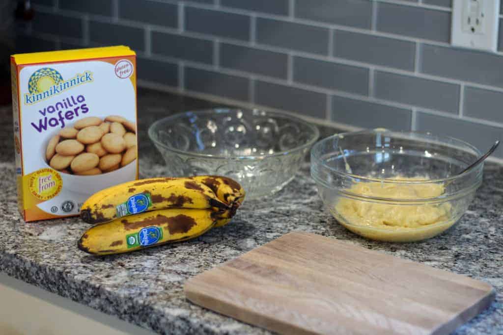 A box of Kinnikinnick's vanilla wafers, three bananas, a bowl of pudding, and crystal bowl sitting on the countertop with a wooden cutting board.