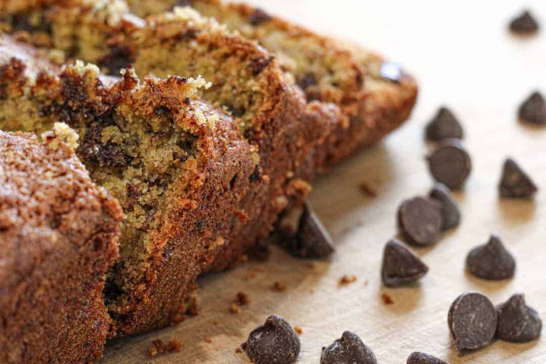 gluten and dairy free chocolate chip banana bread on a wooden cutting board with a few slices cut and leaning forward. Chocolate chips scattered on the cutting board