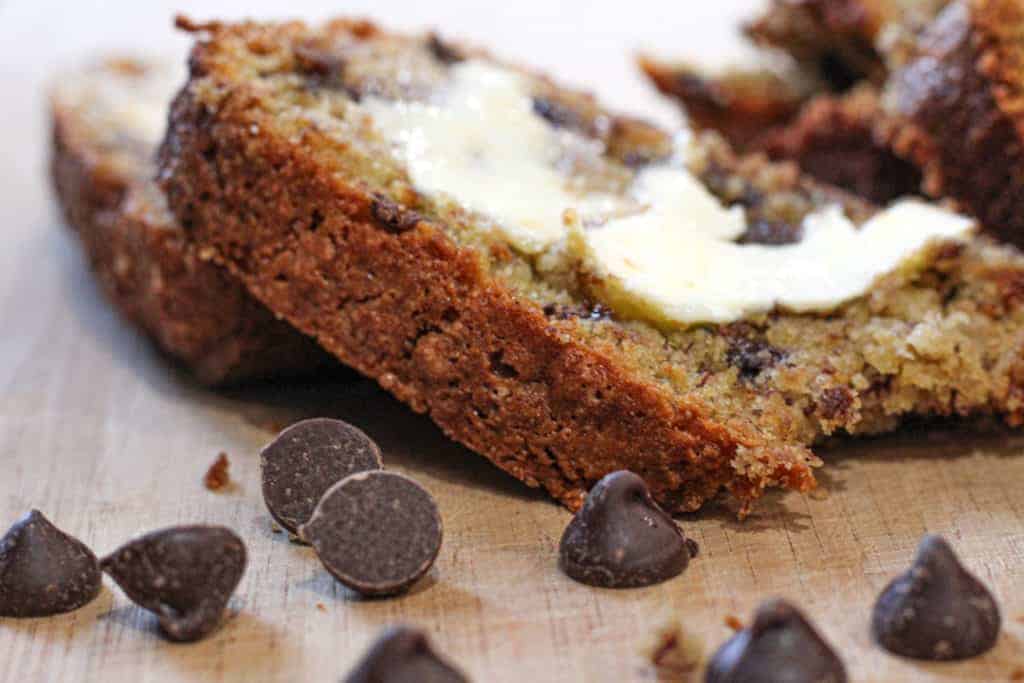 two slices of gluten and dairy free chocolate chip banana bread stacked on top of each other on a wooden cutting board. Chocolate chips are scattered on the board. 