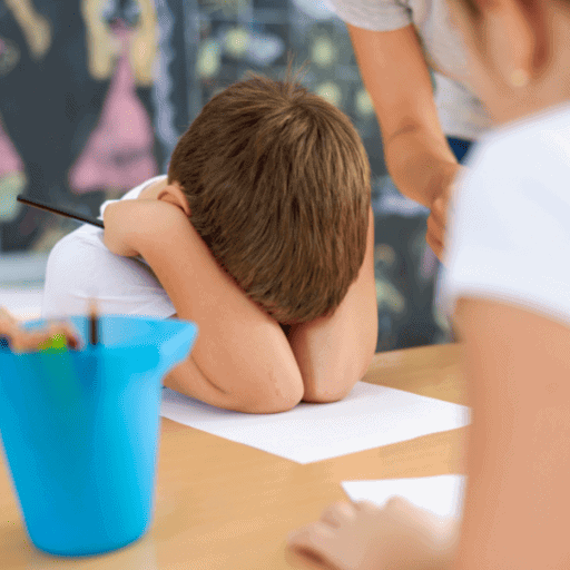 little boy at school with his head down and his arms and hands covering his face and ears ontop of his paper