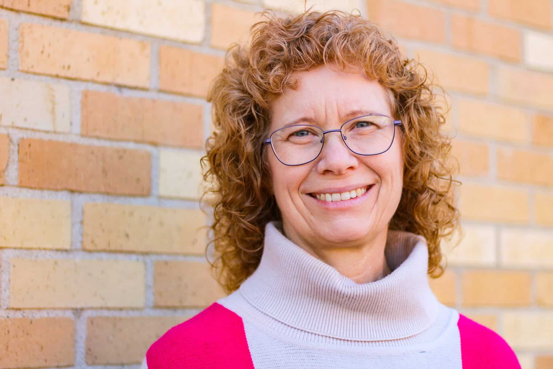 pictures of Cheri Moore wearing glasses standing by a red brick wall wearing a sweater with a cream turtleneck and dark red wide block or red going down.