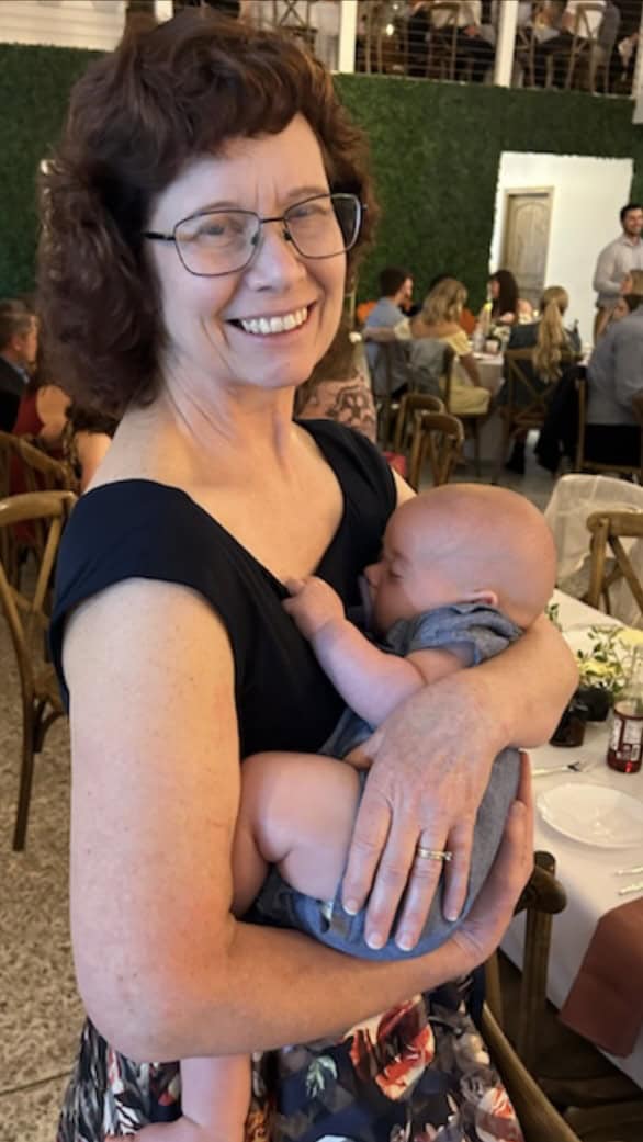 A wedding celebration enjoying conversation at dinner. I am standing up holding my three month old sleeping grandson who is holding onto my dress. I am wearing a navy blue sleeveless dress. 